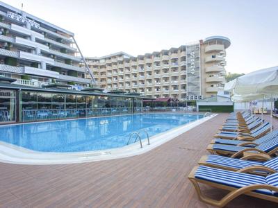 Moderner Außenpool mit Sonnenliegen auf Holzterrasse vor mehrstöckigem Hotelgebäude bei Tageslicht.
