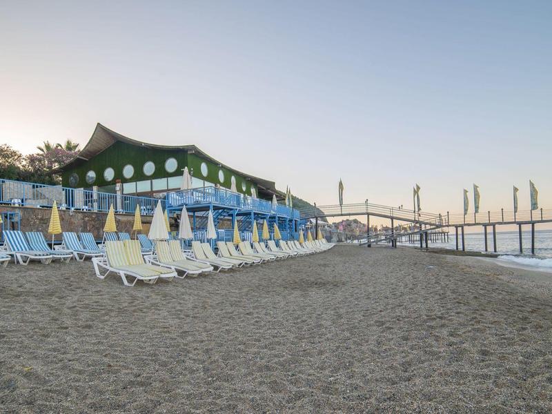 Spiaggia con lettini, edificio verde e ponte sullo sfondo al tramonto.
