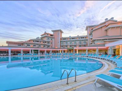 Großer Außenpool mit blauen Liegestühlen vor mehrstöckigem Hotelgebäude bei blauem Himmel.