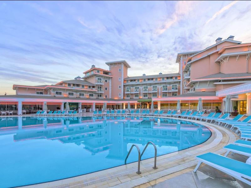 Großer Außenpool mit blauen Liegestühlen vor mehrstöckigem Hotelgebäude bei blauem Himmel.