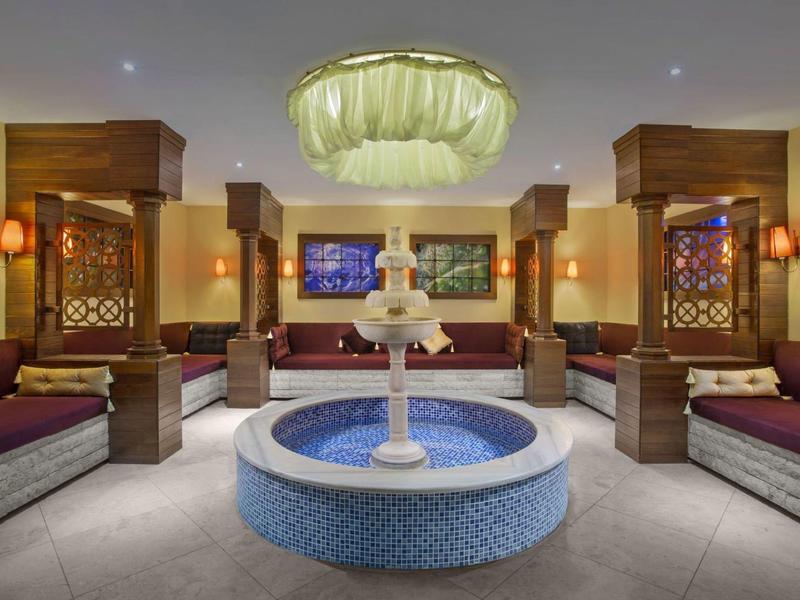 Großer Raum mit rundem Springbrunnen, blauer Mosaikbasis, Säulen und gepolsterten Sitzbänken.