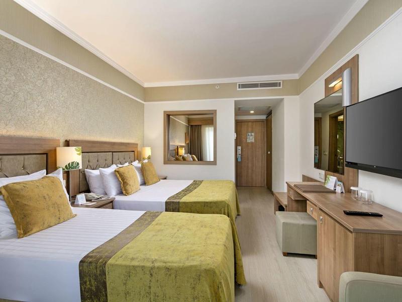 Hotelzimmer mit zwei Betten, grünen Decken, Holzmöbeln und einem großen Flachbildfernseher.