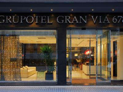 Eingang des Grupotel Gran Via 678 mit großen Glasfenstern und warm beleuchtetem Inneren.