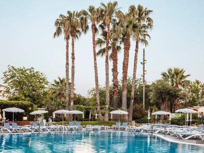 Großer Pool mit blauer Wasseroberfläche, umgeben von Liegestühlen, Palmen und Sonnenschirmen bei klarem Himmel.