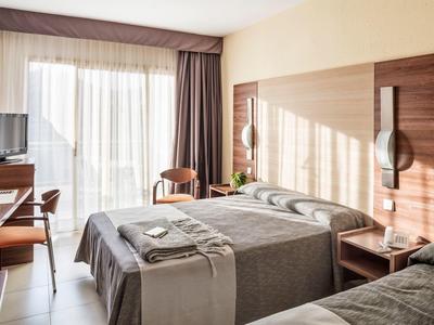 Camera d'albergo luminosa con due letti, mobili in legno e ampia finestra con tende.