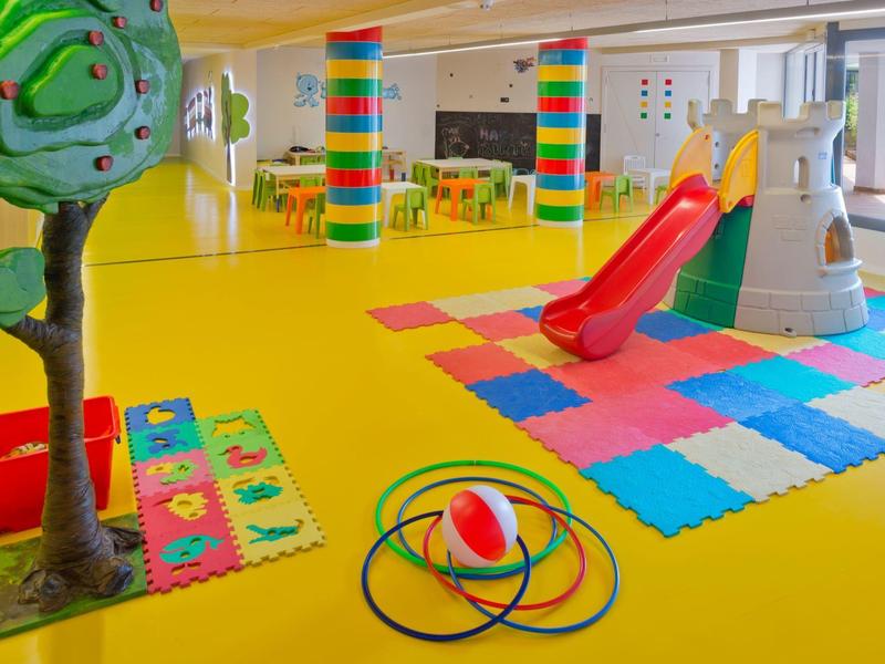 Area giochi interna colorata con scivolo, albero giocattolo, tappetini in schiuma e gioco ad anelli su pavimento giallo.
