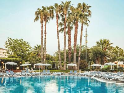 Großer Hotelpool mit Liegestühlen und Palmen im Hintergrund bei klarem Himmel.