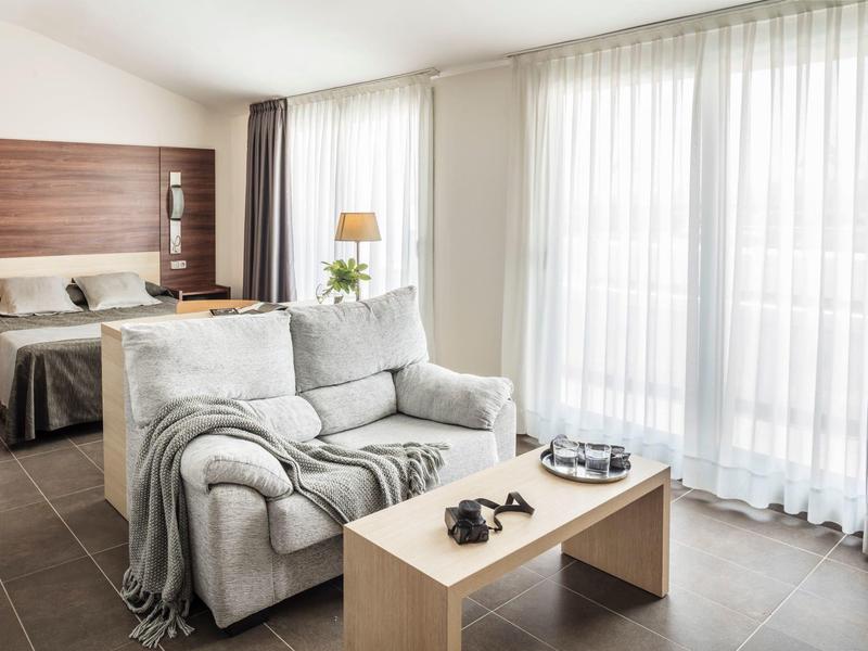 Cozy hotel room with bed, gray sofa, wooden coffee table, and large window with sheer curtains.