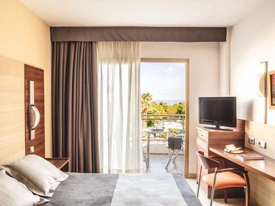 Hotel room with bed, desk, TV, and balcony view of trees and water.