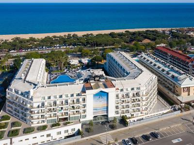 Großes weißes Hotel mit Pool nahe Strand und blauem Meer, viele Balkone und Straßen im Vordergrund.