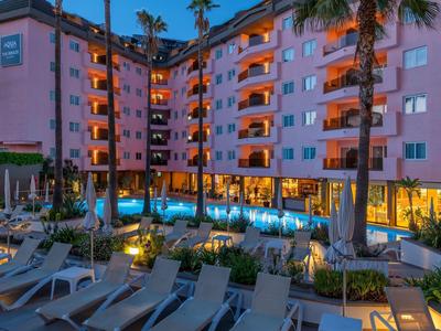 Mehrstöckiges Hotel mit beleuchtetem Pool, Palmen und Liegestühlen am Abend.