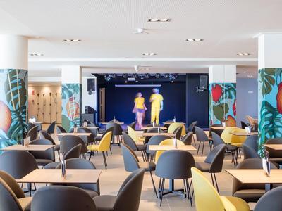 Moderner Veranstaltungsraum mit bunten Säulen, grauen Sesseln, gelben Sofas und Bühne im Hintergrund.