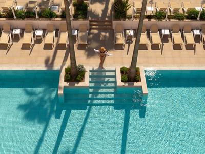 Piscina di un hotel soleggiata con acqua azzurra limpida, palme e file di lettini sul terrazzo beige.