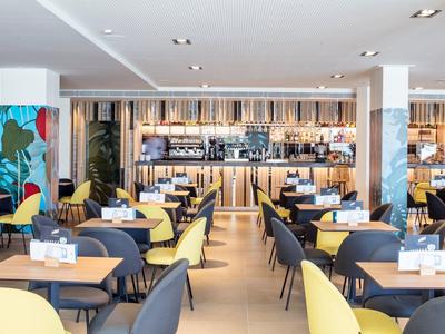 Modernes Restaurant mit gelben und grauen Stühlen, Holztischen und einer Bar im Hintergrund.