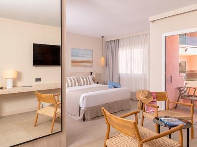 Helles Hotelzimmer mit Doppelbett, Holzstuhl, Schreibtisch, TV und Balkon mit Balkonmöbeln.