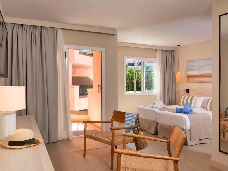 Helles Hotelzimmer mit Holzmöbeln, Bett, Stühlen und großen Fenstern mit Vorhängen.