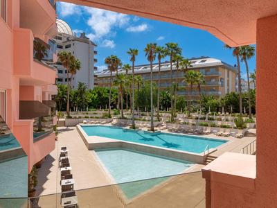 Piscina esterna dell'hotel con palme, circondata da edifici rosa sotto un cielo blu.