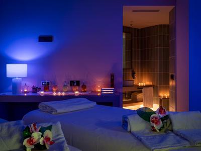 Slabě osvětlená spa místnost s masážními stoly, květinami a svíčkami vytvářející relaxační atmosféru.