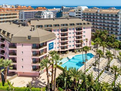 Velký vícepatrový hotel s bazénem a palmami na slunném rekreačním místě.