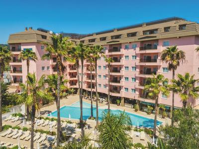 Grande hotel con facciata rosa, palme e una lunga piscina all'aperto sotto un cielo azzurro.