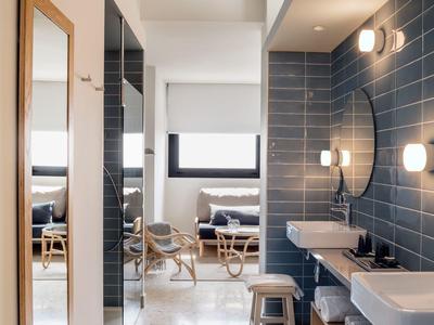 Helles Badezimmer mit blau gekachelter Wand, zwei weißen Waschbecken und Holzregal, Fenster zum Zimmer.