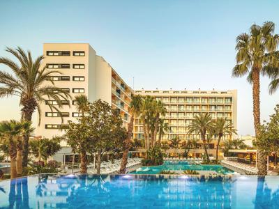 Modern hotel building with large pool and palm trees under clear sky.