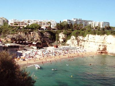 Sunny beach with people, clear water, cliffs, and buildings on the hill in the background.