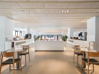 Bright, modern hotel restaurant with wooden chairs, white tables, and large windows.