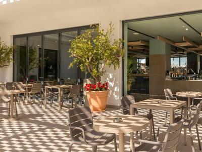 Outdoor seating area with tables and chairs under a shaded patio next to glass windows.