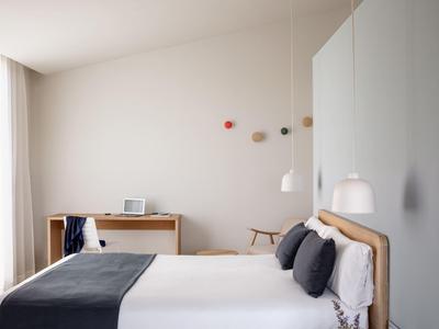 Modern hotel room with wooden bed, white linens, grey cushions, bedside table, desk, and hanging lights.