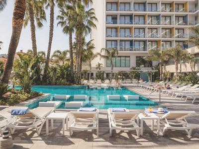 Hotelpoolbereich mit Liegestühlen, Palmen und mehrstöckigem Gebäude im Hintergrund