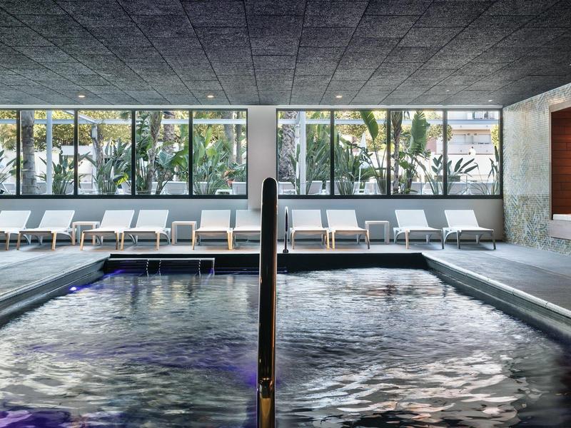 Indoor pool area with lounge chairs and large windows showing palm trees outside.