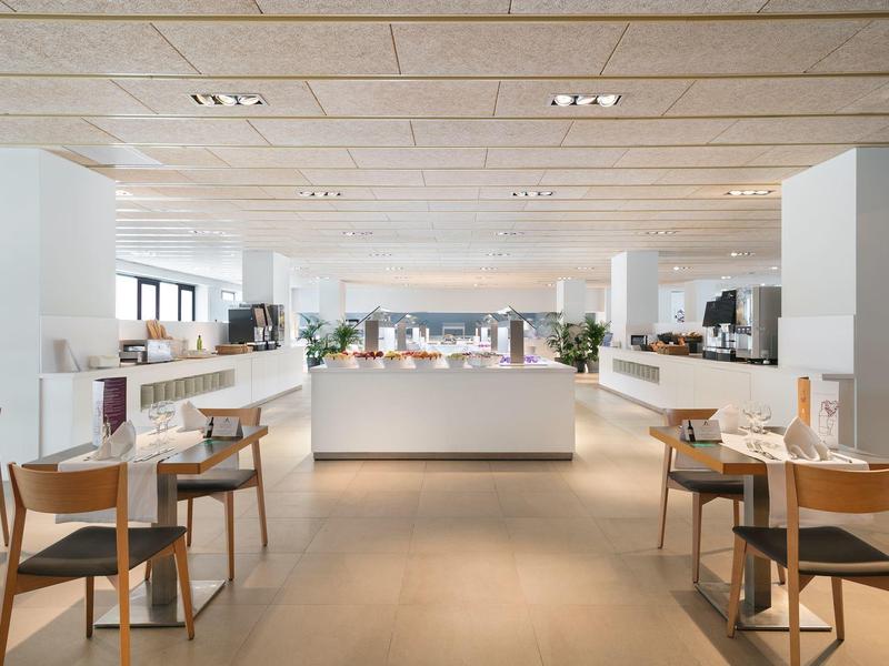 Bright, modern hotel restaurant with wooden chairs, white tables, and large windows.