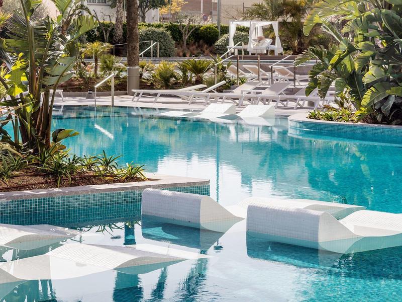 Poolside lounge chairs partially submerged in a clear blue swimming pool surrounded by tropical plants.