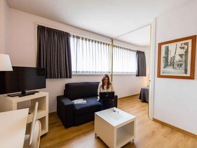 Kleiner Hotelzimmerbereich mit Sofa, Fernseher, Tisch und einer Person mit Laptop.