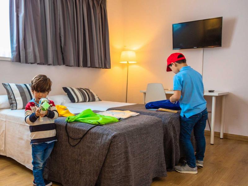 Dva chlapci si hrají v hotelovém pokoji s postelí, televizí a stojací lampou.