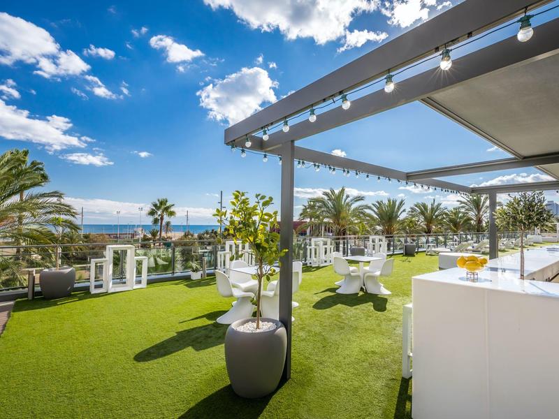 Sunny outdoor terrace with palm trees, white seating, and a bar under a blue sky.