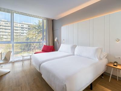 Modernes Hotelzimmer mit zwei weißen Betten, großem Fenster und rotem Sessel neben dem Fenster.