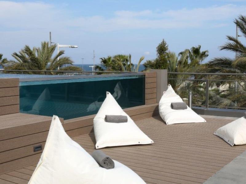 Terrazza sul tetto con panca in legno, quattro cuscini bianchi e vista sulle palme sotto un cielo limpido.