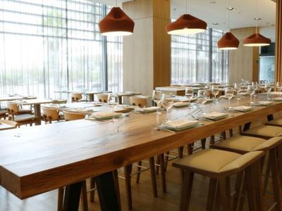 Ristorante luminoso con lunghe tavole di legno e sgabelli, interni moderni e grandi finestre.