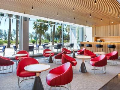 Lounge d'hotel moderno con poltrone rosse, pavimento in legno e grandi finestre con vista sulle palme.