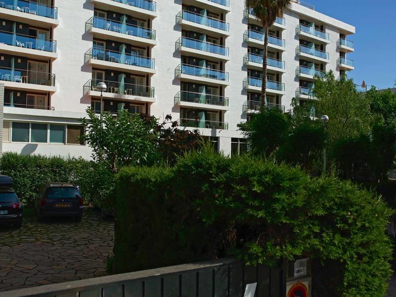 Mehrstöckiges Hotelgebäude mit Balkonen, umgeben von grünen Pflanzen und Parkplätzen.