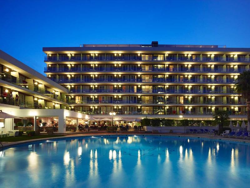 Großes Hotel mit beleuchtetem Pool und mehreren Etagen in der blauen Abenddämmerung.