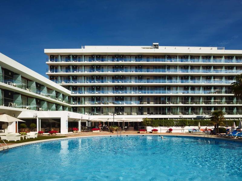Hotelgebäude mit Balkonen neben großem, rundem Pool und blauen Himmel im Hintergrund.
