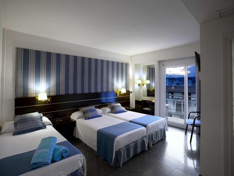 Helles Hotelzimmer mit drei Betten, blauen Akzenten und Balkon mit Blick auf das Meer.