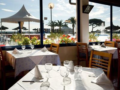 Restaurant mit gedeckten Tischen, Holzstühlen und Ausblick auf Uferpromenade mit Palmen und Segelbooten.