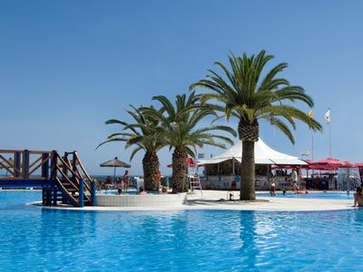 Una piscina con palmeras y un pequeño muelle bajo un cielo despejado, junto a sombrillas y asientos.