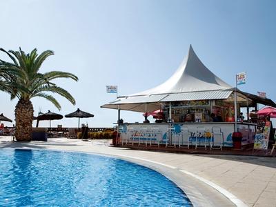 Piscina con un bar y sombrillas junto a una palmera bajo cielo despejado en la playa.