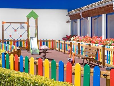 Área de juegos colorida con tobogán, columpios y bancos, rodeada de una cerca y flores.