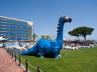 Figura grande de dinosaurio azul en el césped del hotel junto a tumbonas y un edificio hotelero.